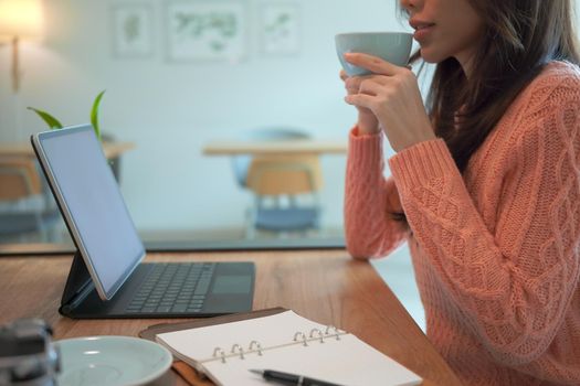 Young asian woman in warm sweater drinking hot coffee and using computer tablet at home.