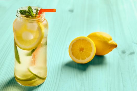 lemons on a turquoise wooden table next to a lemonade in a glass jar, the jug contains water, slices of lemon and lime, ice, mint leaves and a cane to drink