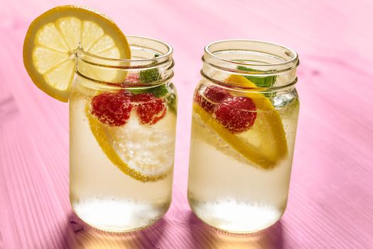 two glass jars with cold water, slices of lemon, mint and red berries, illuminated by sunlight on a pink wooden table with some pieces of citrus, summer refreshments background, copy space