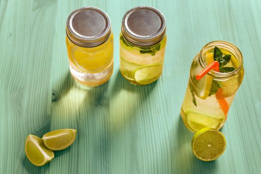 top view of three refreshing lemonades for summer in glass jars, are on a turquoise wooden table and illuminated by sunlight, there are pieces of lemon, lime and mint leaves. Image with copy space
