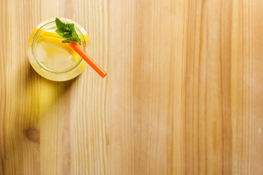 Flat lay of a glass jug with lemonade on a wooden table, the glass contains water, lemon, mint, ice and a straw.