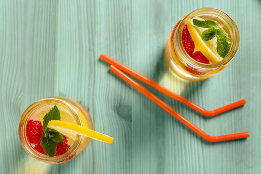 flat lay of two drink canes and two glass jars with refreshing cold lemonade water, lemon, red berries and mint on a green wooden table lit by sunlight, Summer citrus refreshment background Copy space