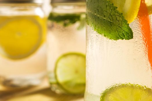 close-up of a glass jar with ice water, lemon, lime and mint leaves, it is on a wooden table and illuminated by sunlight, in the background and out of focus are other lemonades