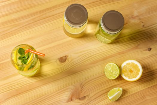 Flat Lay of lemonade with mint and a cane to drink in a glass jar illuminated by sunlight, is on a wooden table, there are also two other jars with lid, some pieces of lemon and lime and copy space