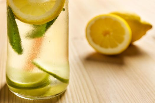 Close-up of the bottom of a glass jar with lemonade on a wooden table and some unfocused lemons in the background. The jar contains water, slices of lime and lemon, ice and mint leaves