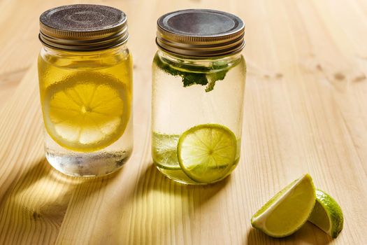 cold summer refreshments with slices of citrus and mint leaves, are in glass jars with lids and illuminated by sunlight, on a wooden table, there are also lemon wedges