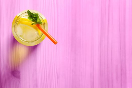 Flat lay of a glass jug with lemonade on a pink wooden table, the glass contains water, lemon, mint, ice and a straw.