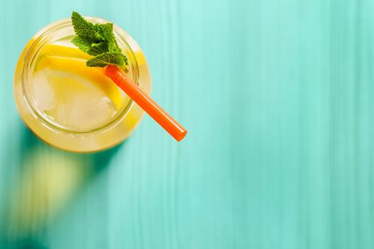 Flat lay of a glass jug with lemonade on a turquoise wooden table, the glass contains water, lemon, mint, ice and a straw.