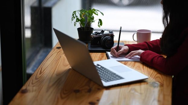 Cropped shot young female freelancer using laptop computer and making notes on notebook.