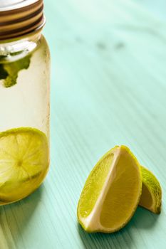 vertical photo of a glass jar with a lid full of lemonade next to some slices of lime, the glass is illuminated by sunlight and on a turquoise wooden table