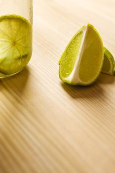 vertical photo with copy space of some slices of lime next to a glass jar with lemonade, this is illuminated by sunlight and on a wooden table