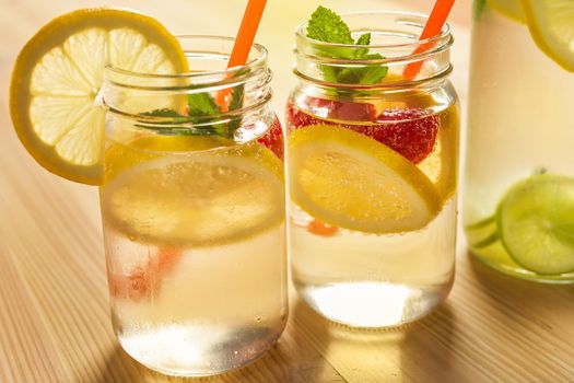 two glass jars illuminated by sunlight with refreshing cold lemonade water, lemon slices, red berries, mint leaves and drinking canes on a wooden table. Summer refreshment background