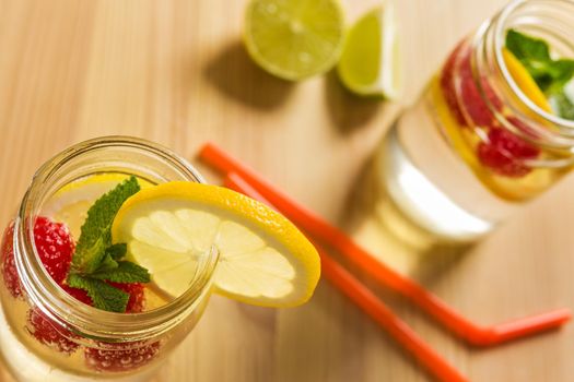 top view, glass jar illuminated by sunlight with refreshing cold lemonade water, slices of lemon, red berries and mint on a unfocused wooden table with canes and pieces of citrus. Summer refreshment