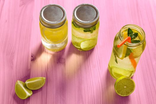 top view of three refreshing lemonades for summer in glass jars, are on a pink wooden table and illuminated by sunlight, there are pieces of lemon, lime and mint leaves. Image with copy space