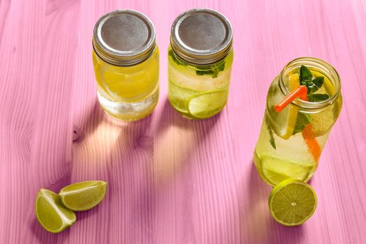 top view of three refreshing lemonades for summer in glass jars, are on a pink wooden table and illuminated by sunlight, there are pieces of lemon, lime and mint leaves. Image with copy space