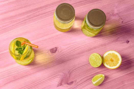 Flat Lay of lemonade with mint and a cane to drink in a glass jar illuminated by sunlight, is on a pink wooden table with two other jars with lid, some pieces of lemon and lime and copy space