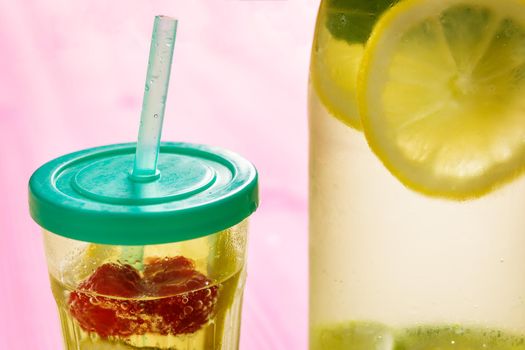 glass with lid and cane and a bottle, with cold water and slices of lemon, lime, berries and mint, are illuminated by sunlight on a wooden pink table with some pieces of citrus