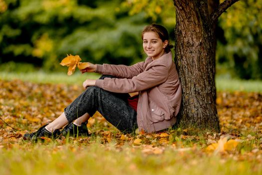 A teenage girl sits in a park under an autumn tree