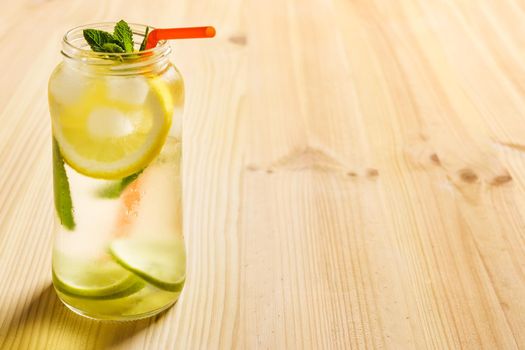 horizontal photo of a lemonade in a glass jar on a wooden table, the jug contains water, slices of lemon and lime, ice, mint leaves and a cane to drink