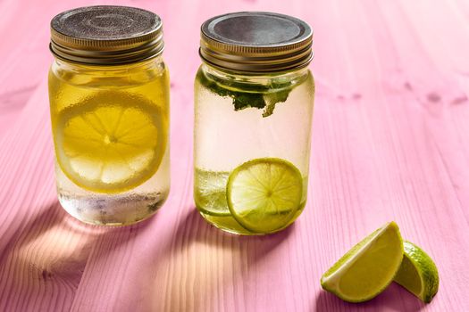 cold summer refreshments with slices of citrus and mint leaves, are in glass jars with lids and illuminated by sunlight, on a pink wooden table, there are also lemon wedges