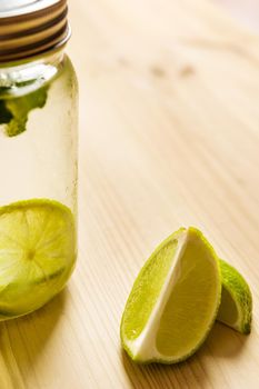 vertical photo of a glass jar with a lid full of lemonade next to some slices of lime, the glass is illuminated by sunlight and on a wooden table