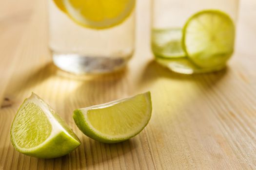 two slices of lemon on a wooden table with homemade citrus sodas unfocused in the background and illuminated by sunlight, the glasses are filled with water with slices of lemon and lime