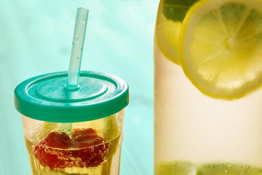 glass with lid and cane and a bottle, with cold water and slices of lemon, lime, berries and mint, are illuminated by sunlight on a wooden green table with some pieces of citrus