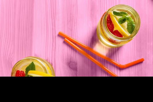 flat lay of two drink canes and two glass jars with refreshing cold lemonade water, lemon, red berries and mint on a pink wooden table lit by sunlight, Summer citrus refreshment background, Copy space