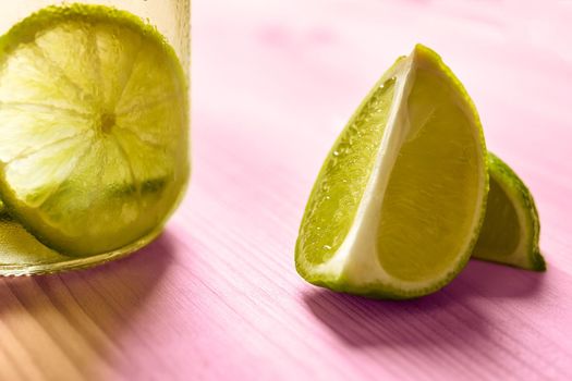 horizontal photo of some slices of lime next to a glass jar with lemonade, this one is illuminated by sunlight and on a pink wooden table