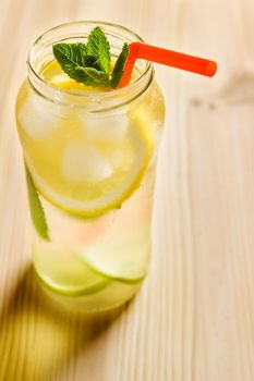 vertical photo of a lemonade in a glass jar on a wooden table, the jug contains water, slices of lemon and lime, ice, mint leaves and a cane to drink