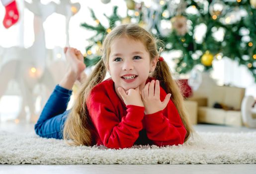 Child girl in Christmas time lying on fluffy carpet and looking at camera in room with decorated tree and gifts. Pretty kid at home in New Year holidays