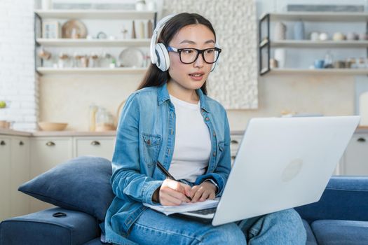 Young beautiful Asian woman studying at home remotely, female student listening to online course taking notes, sitting on sofa at home in living room using headphones and laptop