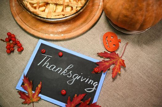 Flat lay. Chalk lettering Thanksgiving Day on a black board with fallen dry autumn maple leaves, ripe pumpkin and festive apple pie with flaky crispy crust on a table with sackcloth tablecloth