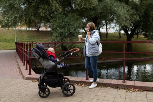 adult mother walks with a baby in a pram in a city park talking on a mobile phone.