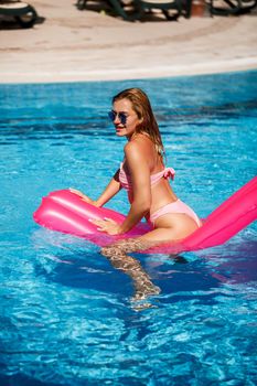 Sexy female model in sunglasses resting and sunbathing on a mattress in the pool. Woman in a pink bikini swimsuit floating on an inflatable pink mattress. spf and sunscreen