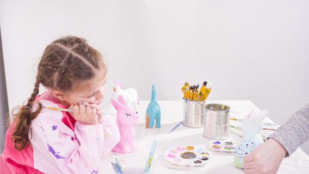 Little girl painting paper mache figurines with acrylic paint for her homeschooling art project.