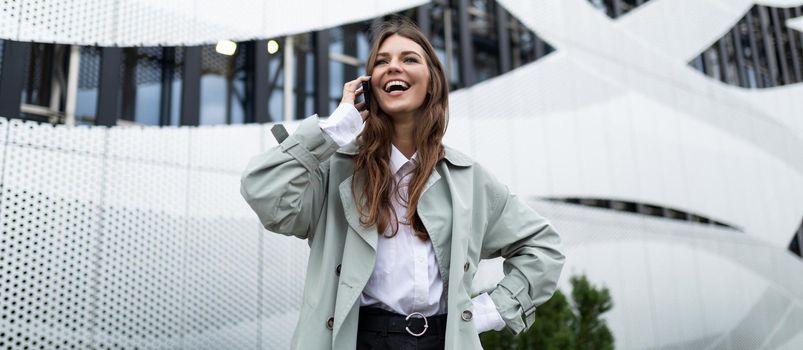 a young woman speaks on a mobile phone with a wide smile on her face against the background of an aluminum facade of an office building.