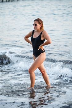 Portrait of a beautiful woman in a black swimsuit with blond hair posing on the beach by the sea. Young woman walking on the beach