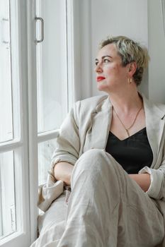 A middle-aged woman in a beige suit and black tank top sits mysteriously and looks out the window on the windowsill. Green trees outside.