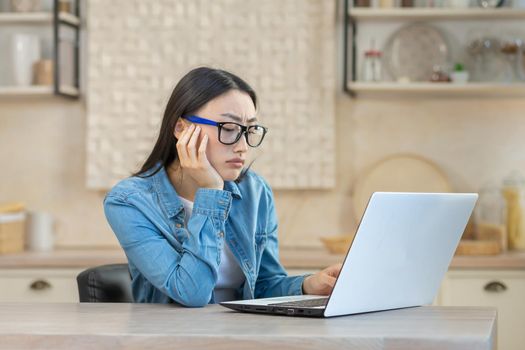 Sad and upset woman sitting at home in the kitchen, asian woman with laptop working online remotely, depressed student,