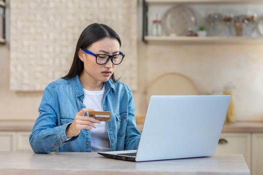 Upset and sad woman in depression trying to make online purchase in online store, Asian woman sitting at home in kitchen using laptop and bank credit card, unable to make payment online.