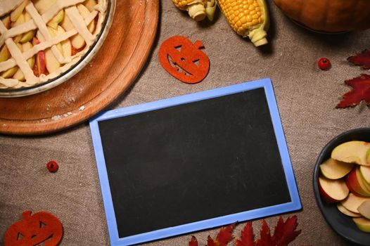 Still life with a black board with copy space for advertising text, maple autumn leaves and partial view of a fresh pumpkin pie for Thanksgiving dinner and harvested corn, on a linen tablecloth