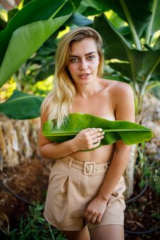 A beautiful young woman stands in a beautiful pose near a banana tree, her little girl covers her breasts with a leaf on a banana plantation.