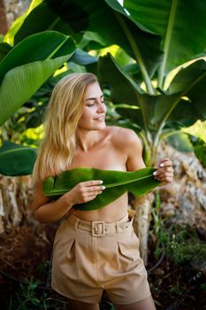 A beautiful young woman stands in a beautiful pose near a banana tree, her little girl covers her breasts with a leaf on a banana plantation.