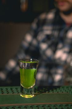 Closeup of shot glass with layered drink Green Mexican on bar counter against blurred background of plaid shirt of bartender. Concept of making cocktails