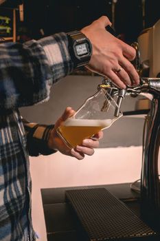 Male bartender in plaid shirt standing behind bar counter filling glass with white unfiltered beer from tap in pub, cropped shot