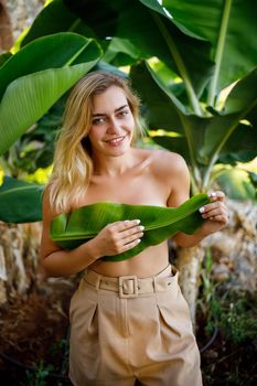 A beautiful young woman stands in a beautiful pose near a banana tree, her little girl covers her breasts with a leaf on a banana plantation.