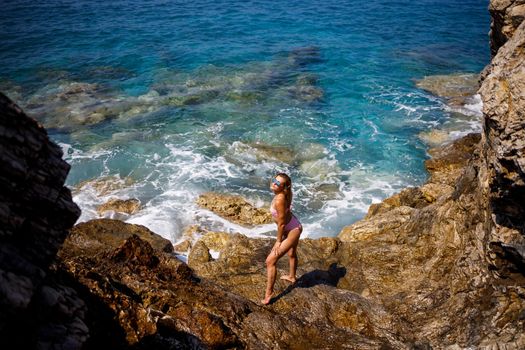 Young beautiful woman in a swimsuit stands on a rocky beach of the Mediterranean Sea. The concept of sea recreation. Selective focus