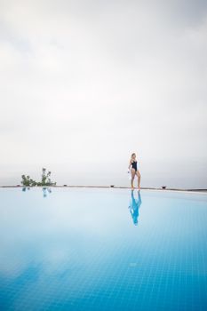 Beautiful woman in black swimsuit posing near outdoor pool with sea view