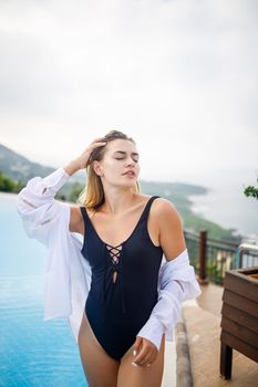 Beautiful woman in black swimsuit posing near outdoor pool with sea view